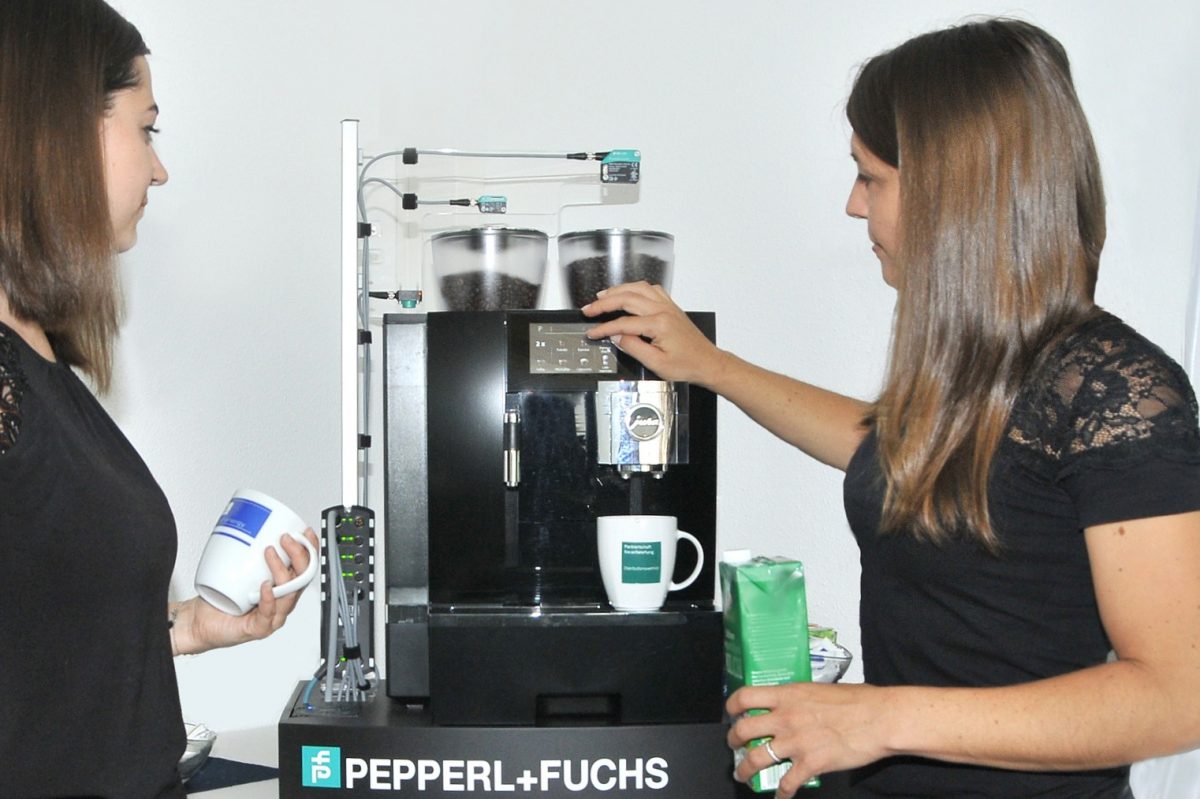 Two women are standing to the left and right of the smart coffee machine with sensors and cloud connectivity. The woman on the left is holding an empty coffee cup. The woman to the right of the coffee machine is pressing one of the buttons with her right hand and holding a carton of milk in her left hand. There is an empty cup under the coffee dispenser.
