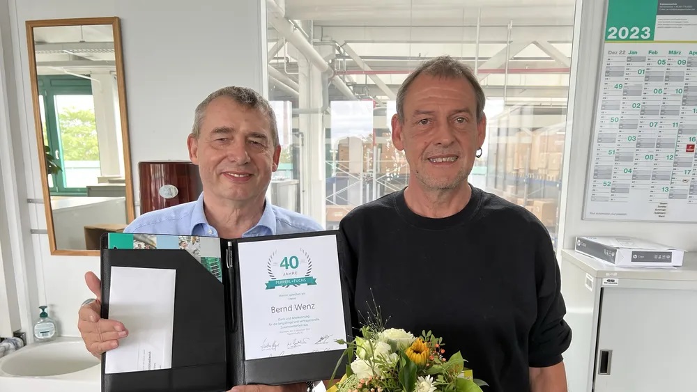 Andreas presents Bernd with the certificate for his 40 years of service. Andreas holds the certificate into the camera and Bernd holds a bouquet of flowers in his hand. They both look into the camera.