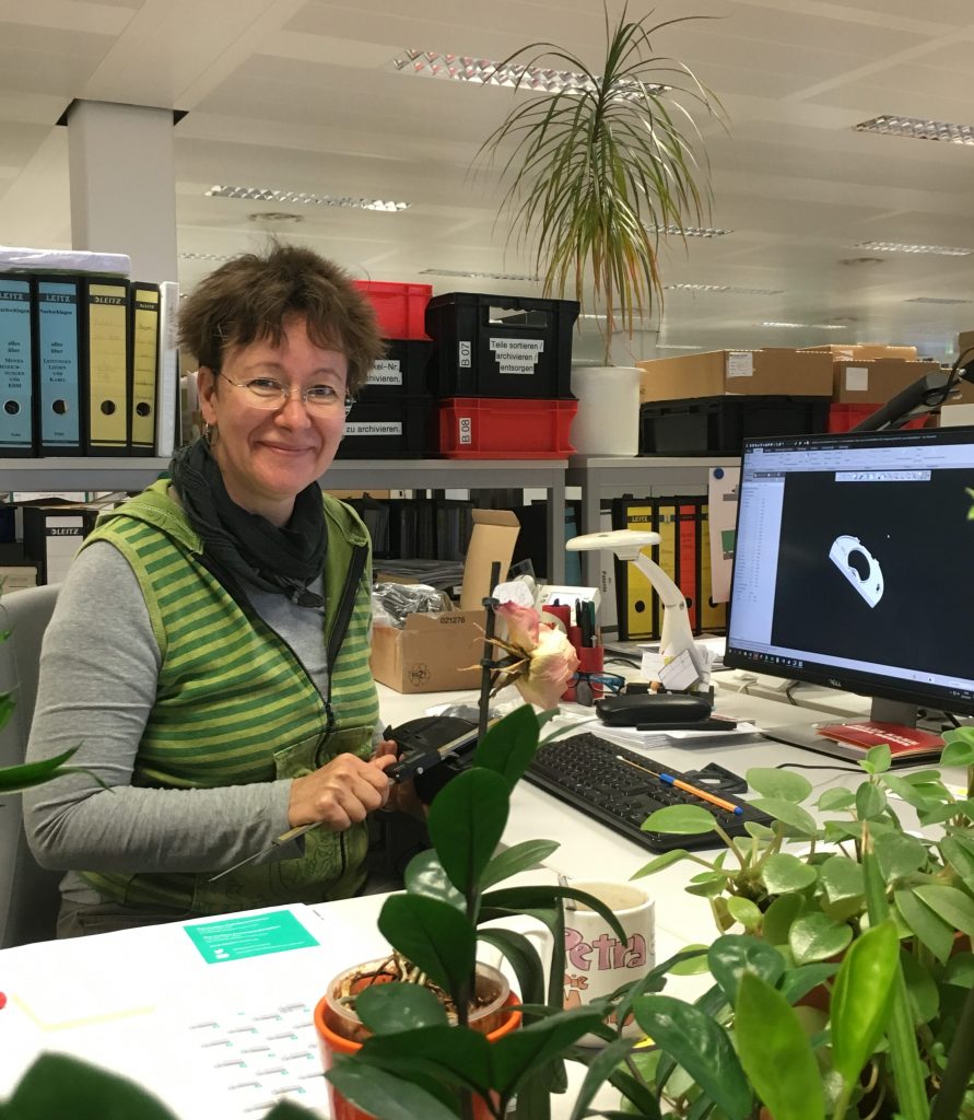 Konstrukteurin Petra an ihrem Arbeitsplatz. Sie sitzt an einem Eckschreibtisch. Neben PC, Tastatur und anderen Büromaterialien stehen auch verschiedene Pflanzen auf ihrem Tisch. Im Hintergrund steht ein Regal mit vielen Ordnern.