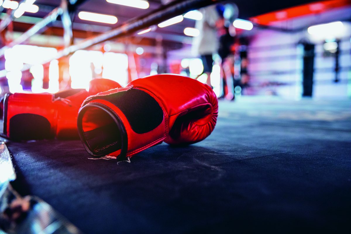Nahaufnahme von roten Boxhandschuhen, die am Rande eines Boxrings liegen. Im Hintergrund sieht man unscharf Boxer beim Training.