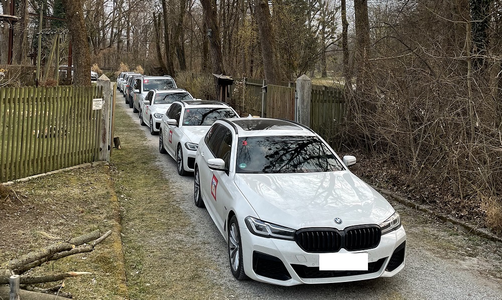 Kolonne von parkenden, weißen BMWs. Sie stehen zwischen eingezäunten Grundstücken auf einem Schotterweg.