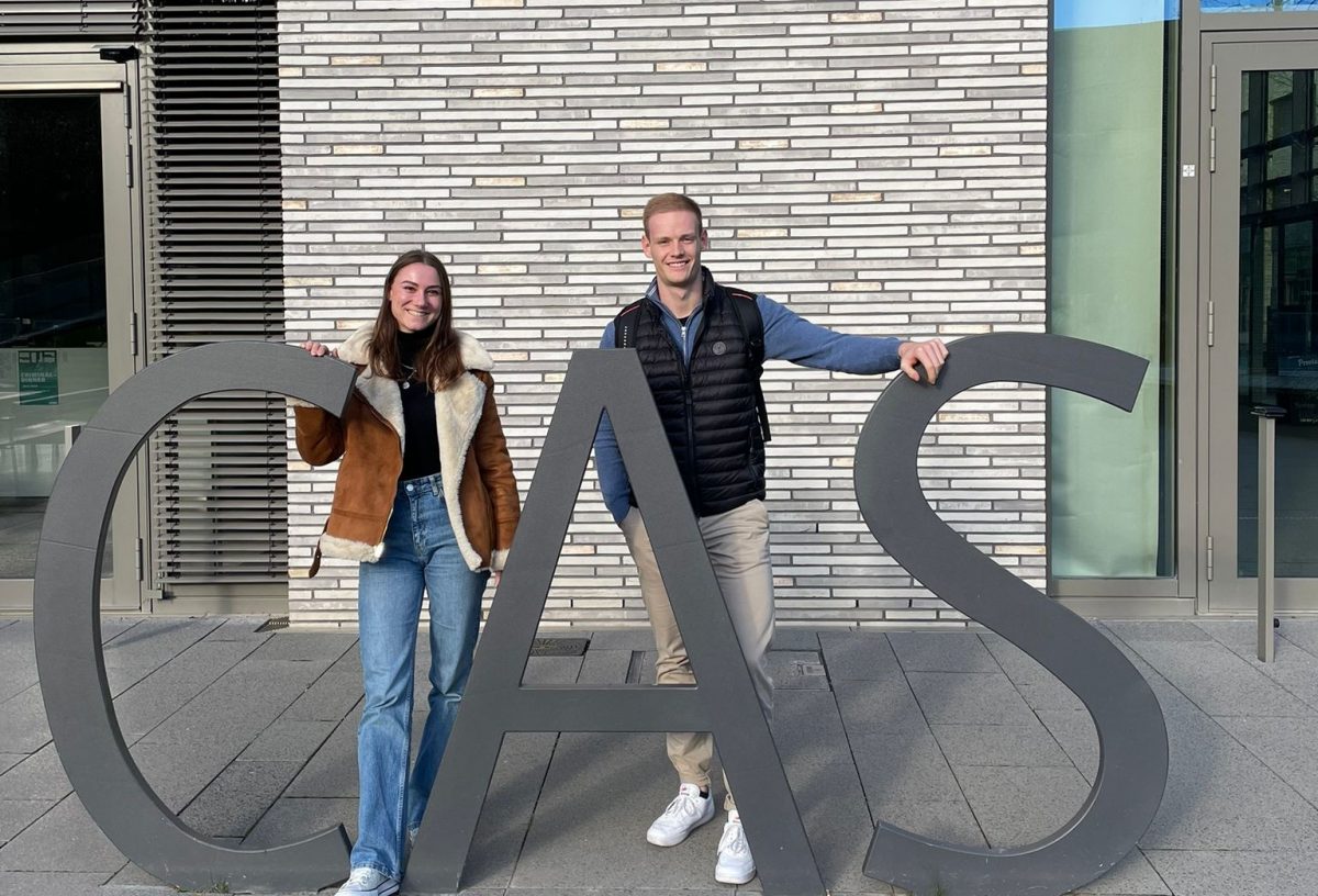 Carina und Max, beides duale Masterstudierende bei Pepperl+Fuchs, lehnen sich an die fast lebensgroßen Metallbuchtaben C A S vor dem Gebäude der DHBW CAS und lächeln in die Kamera.
