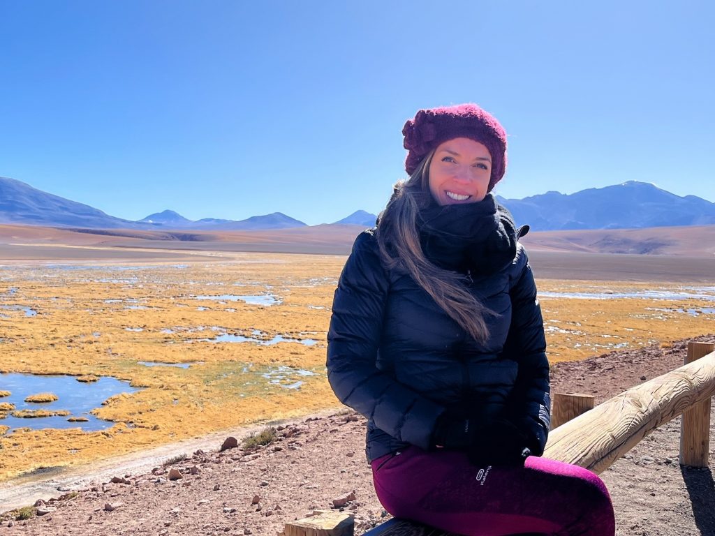 Auf diesem Bild sieht man Carina in der Atacama Wüste in Chile. Sie sitzt auf einer Holzabsperrung und lächelt in die Kamera.