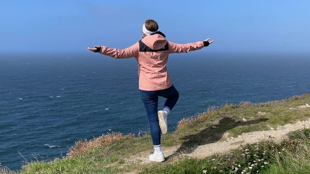 Anna-Lena steht mit dem Rücken zur Kamera auf einem kleinen Trampelpfad. Sie blickt auf das Meer, hat die Arme ausgestreckt und ein Bein angewinkelt.