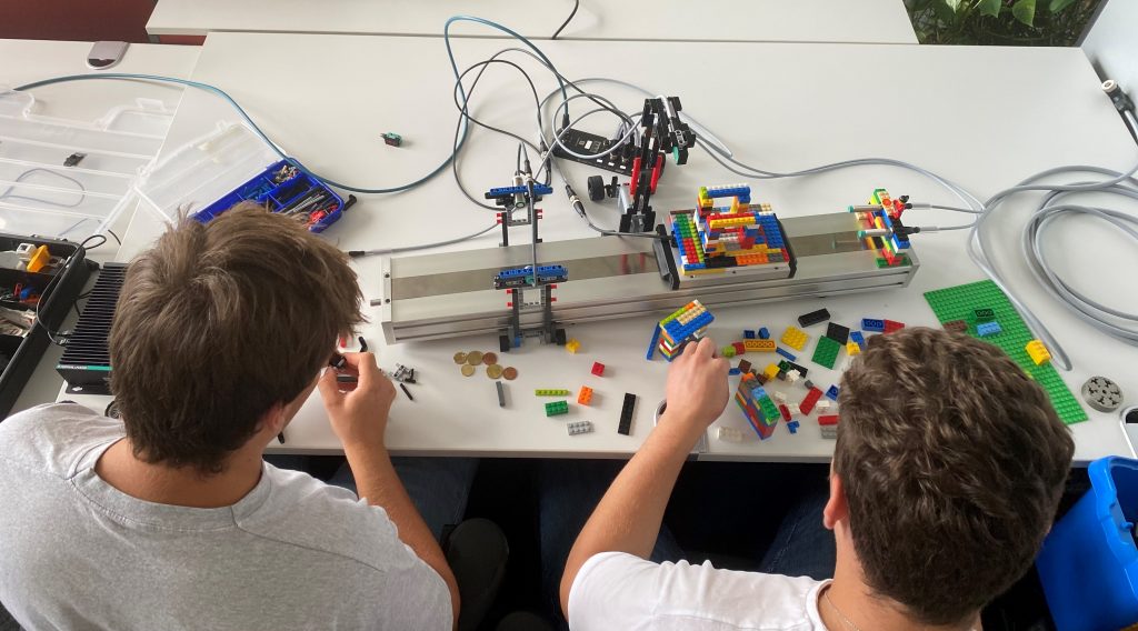 Blick über die Schulter von Aaron und Fabian, die beide das Studium Elektrotechnik bei Pepperl+Fuchs absolvieren. Sie bauen gerade an der Halterung mit Legobausteinen. Vor ihnen liegt die bisherige Konstruktion.