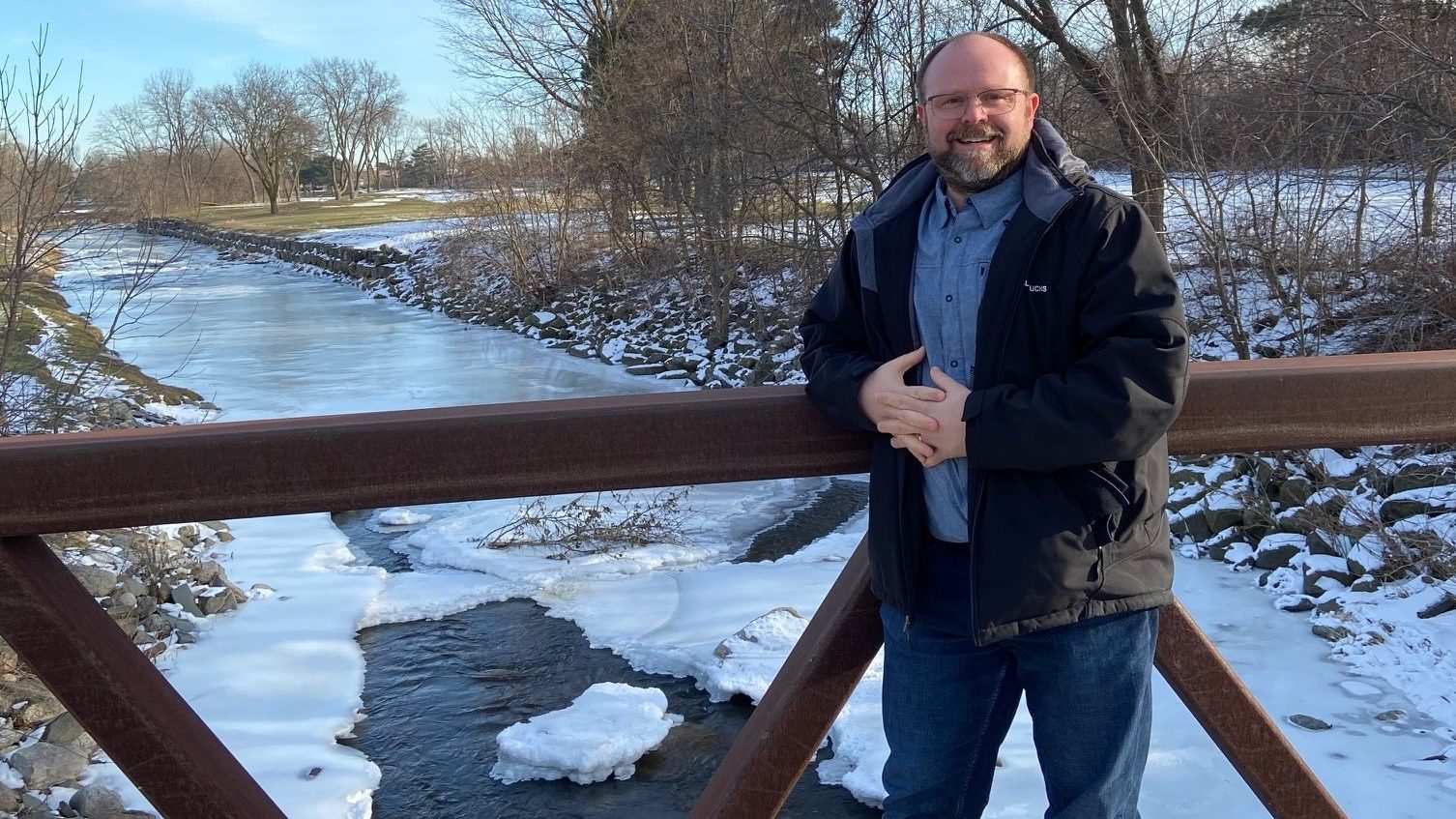 Richard lehnt an einer Brücke, die über einen zum Teil zugefrorenen Fluss führt. Die Bäume sind kahl, es liegt etwas Schnee und die Sonne scheint.