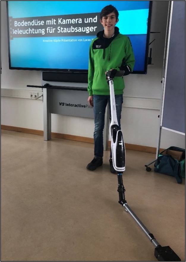 Lucas steht mit dem fertigen Staubsauger vor einem großen Fernseher und lächelt stolz in die Kamera. Auf dem Fernseher ist nochmals sein Projektitel "Bodendüse mit Kamera und Beleuchtung für Staubsauger" zu lesen.