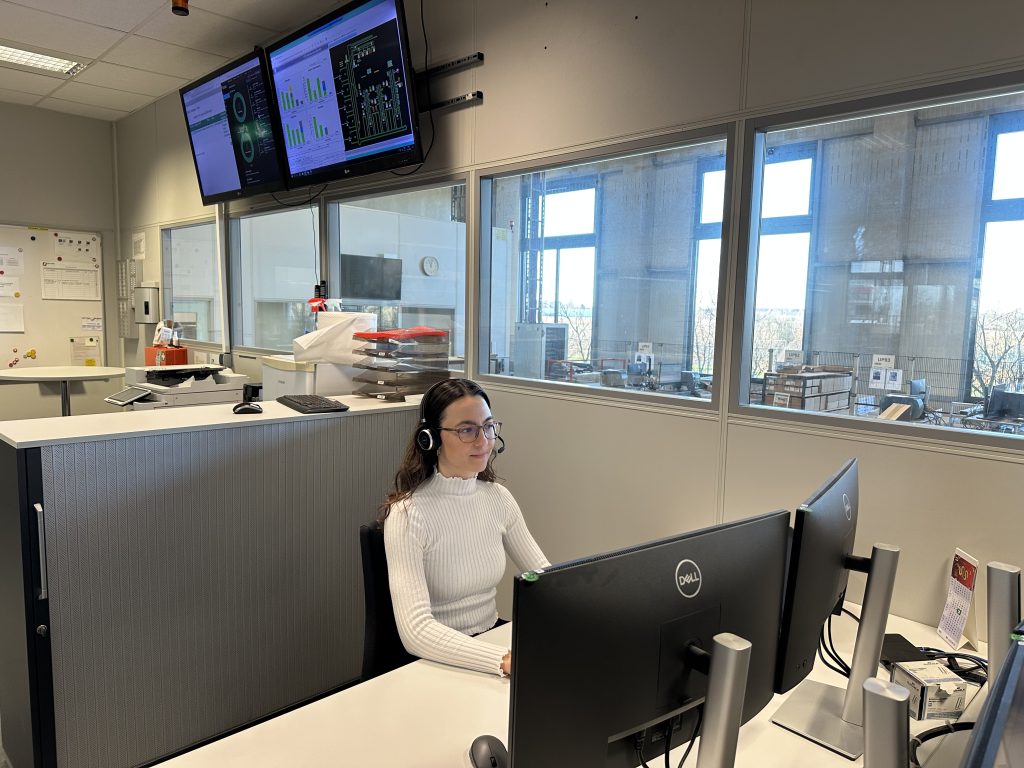 Rebecca ist Projekt- und Prozessingenieurin bei Pepperl+Fuchs. Hier sitzt sie an ihrem Schreibtisch und arbeitet. Im Hintergrund sind weitere Arbeitsplätze zu sehen, sowie zwei Monitore, die an der Wand über ihr hängen.