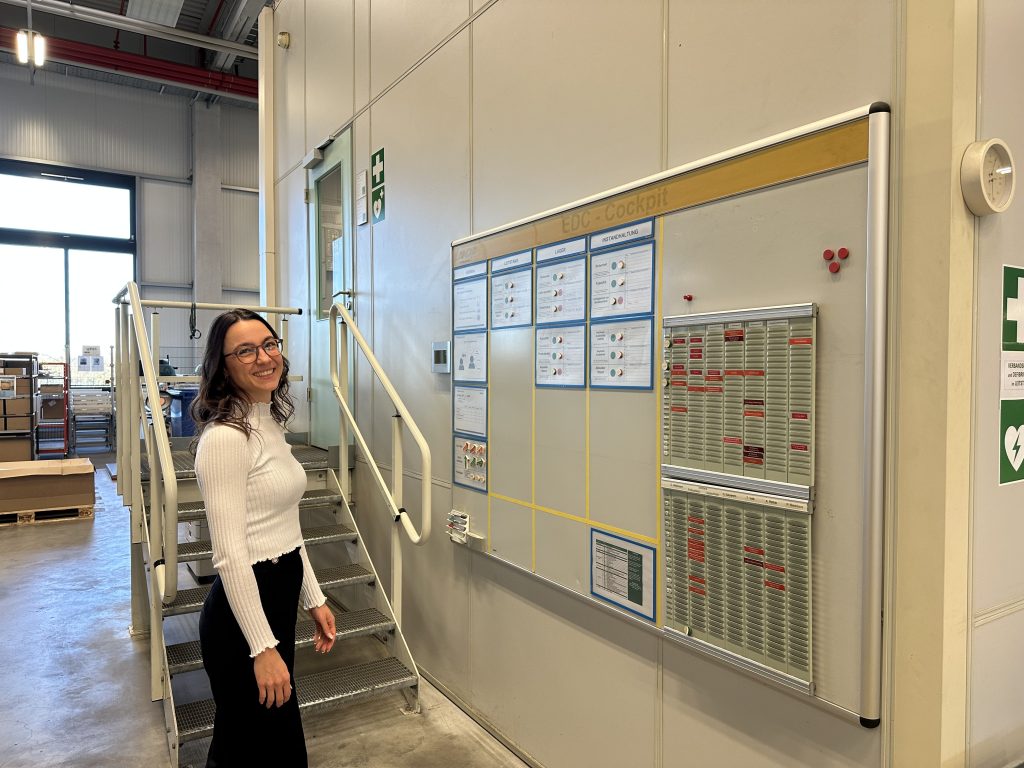 Rebecca auf dem Weg durch die Logistik von Pepperl+Fuchs. Rechts von ihr hängt ein großes Board, welches verschiedene Informationen aufzeigt.