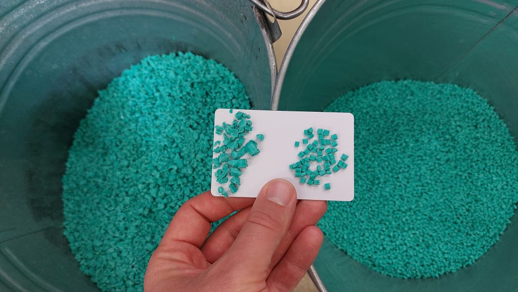 Zwei Tonnen sind zu sehen. In der linken Tonne ist das gemahlene Material (Regrind), in der rechten Tonne das Neugranulat. Eine Hand ist zu sehen, welche einen kleinen, weißen Karton in der Hand hält, auf dem die beiden Materialien nochmals näher nebeneinander zu sehen sind. Das Material hat eine türkise Farbe.