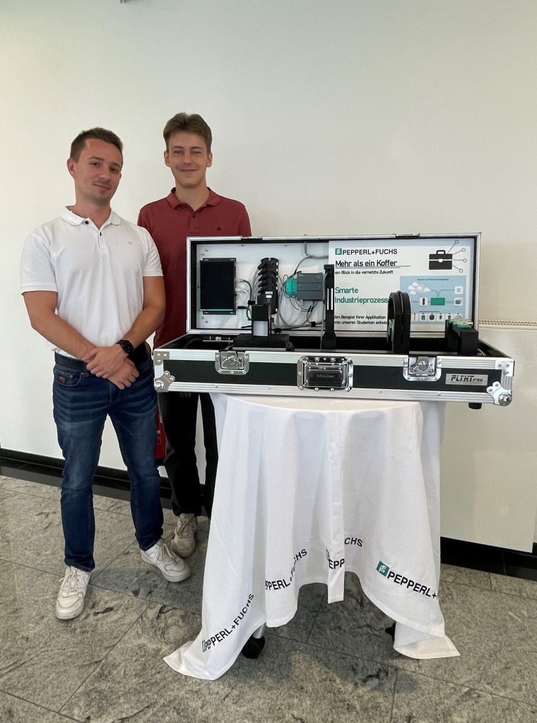 Praxisphasen bei Pepperl+Fuchs: Daniel und David stehen hinter dem geöffneten Kofferexponat, welches auf einem runden Tisch mit einem weißen Tischtuch mit Pepperl+Fuchs Logo steht. Im Koffer sieht man die verbaute Sensorik.