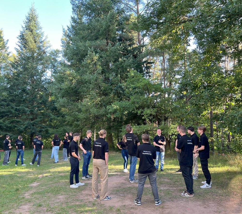 Gruppe junger Menschen steht im Kreis auf einer Wiese und macht ein Kennenlern-Spiel. In der Mitte des Kreises steht ebenfalls eine Person. Sie sind umgeben von hohen Bäumen.