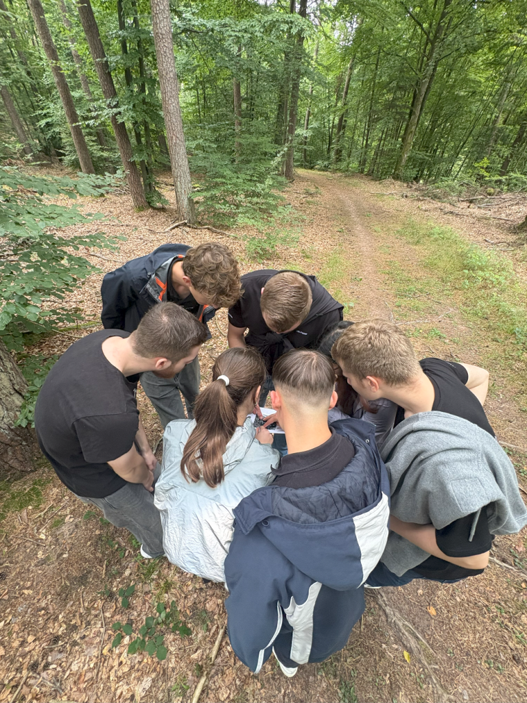 Eine Gruppe von sechs Personen kniet auf einem Waldweg auf dem Boden und beugen sich gemeinsam über ein Stück Papier.