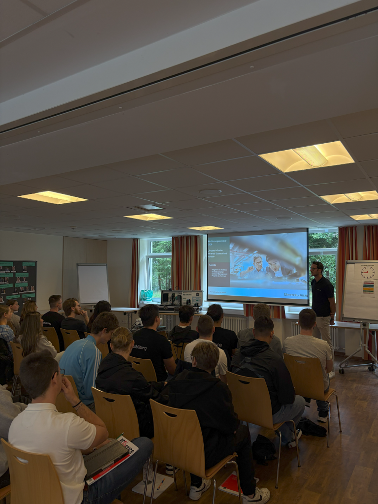 Ausbildungsstart bei Pepperl+Fuchs: Die Auszubildenden und dual Studierenden sitzen in einem Raum und hören einem Vortrag zu. Man sieht mehrere bestuhlte Reihen, auf denen Personen sitzen. Vorne steht ein Mann neben einer Leinwand, auf der das Thema des Vortrags zu sehen ist.